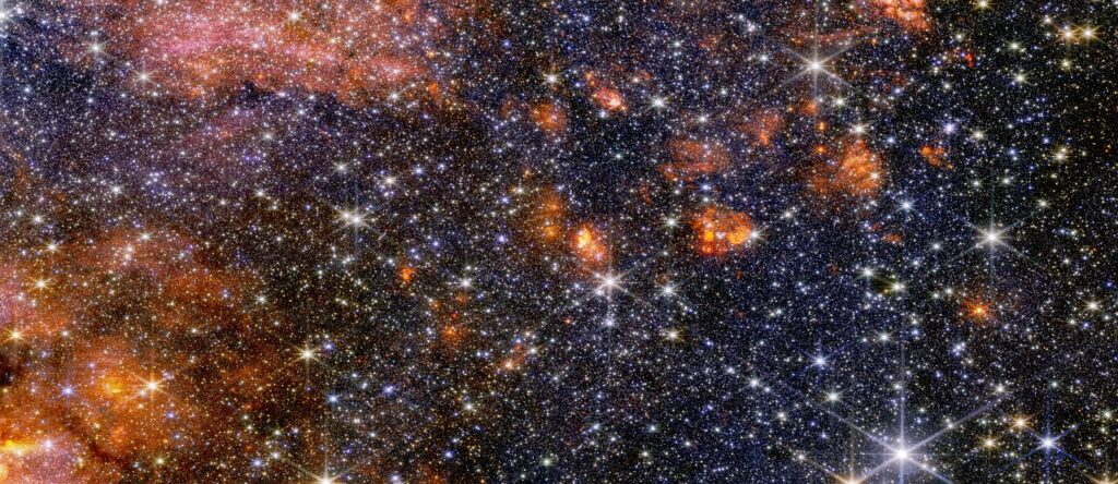 Stars, gas and cosmic dust in the Sagittarius B2 molecular cloud glow in near-infrared light, captured by Webb’s NIRCam (Near-Infrared Camera). In this light, astronomers see more of the region’s diverse, colourful stars, but less of its gas and dust structure. Webb’s instruments each provide astronomers with important information that help build a more complete picture of what is happening in this intriguing portion of the centre of our galaxy. [Image description: A wide view of a region of space filled with stars and clumps of orange clouds.]